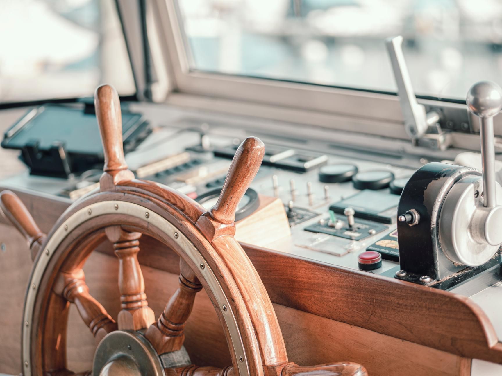 Wooden ship's wheel on a boat, with control panel and window in the background.