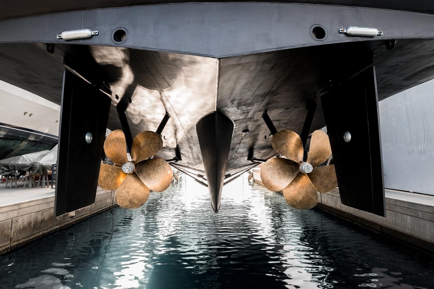 Bronze boat propellers under a large ship's hull in a dry dock, viewed from below.