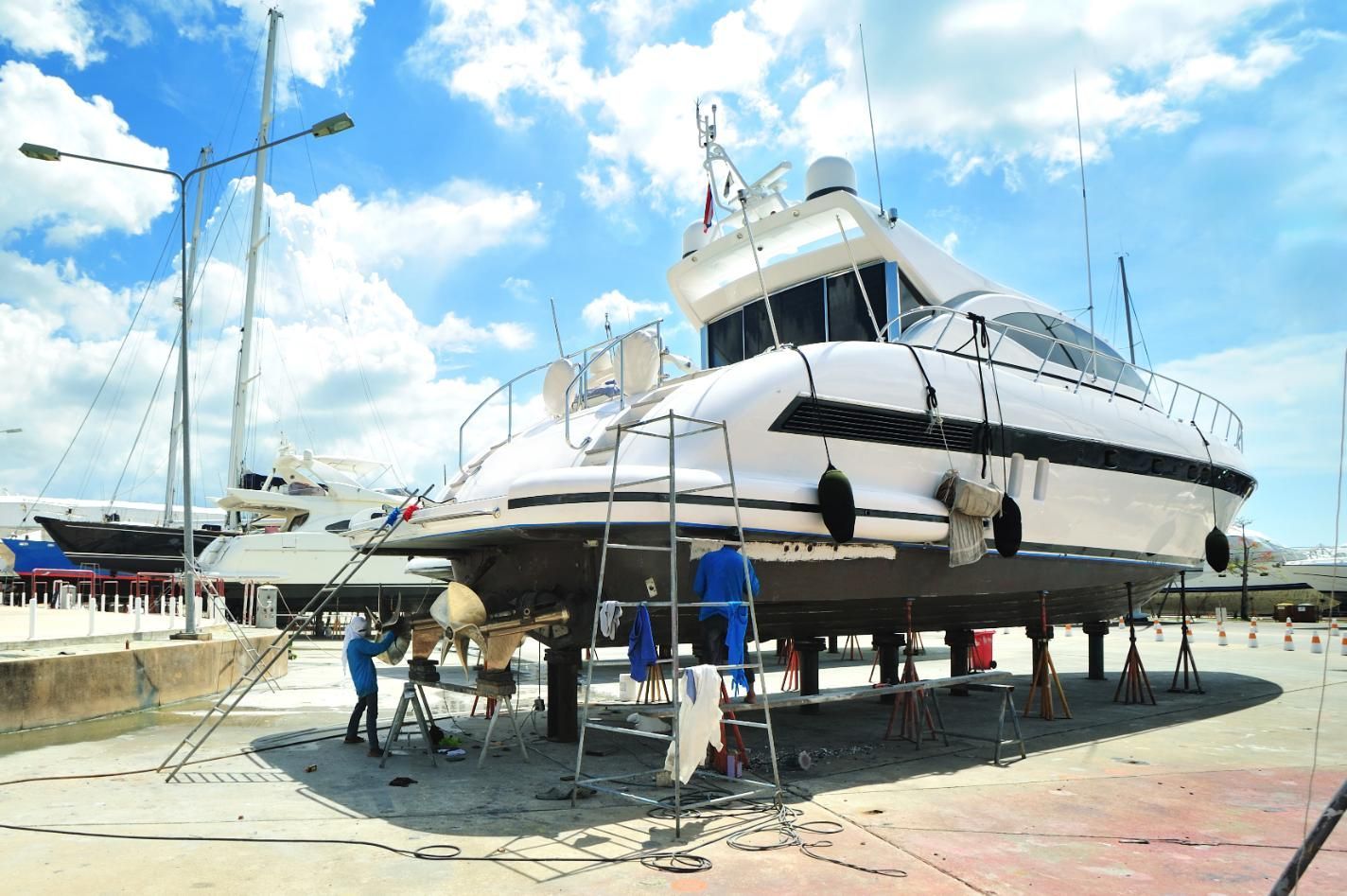 Boat being worked on in a dry dock, with workers, blue sky, and other boats visible.