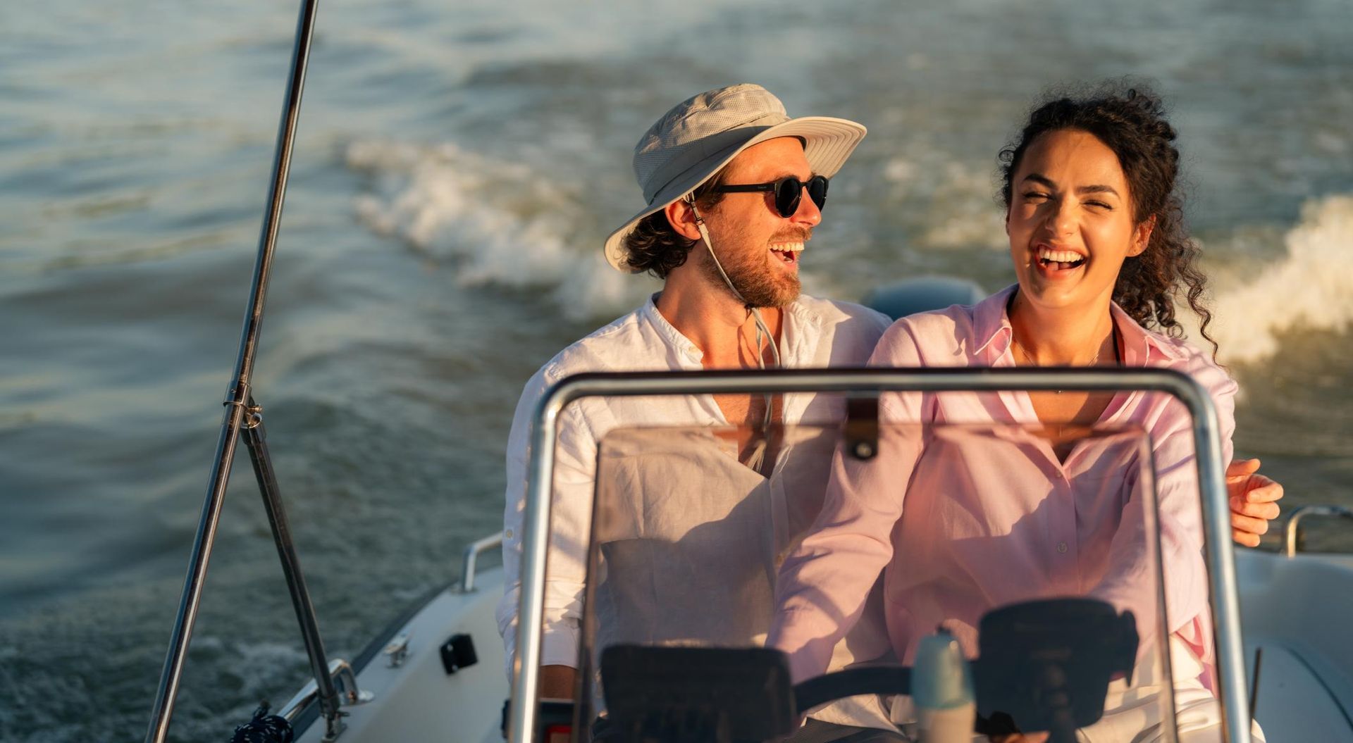 Couple laughing while boating on a sunny day. Man at helm, wearing hat and sunglasses. 