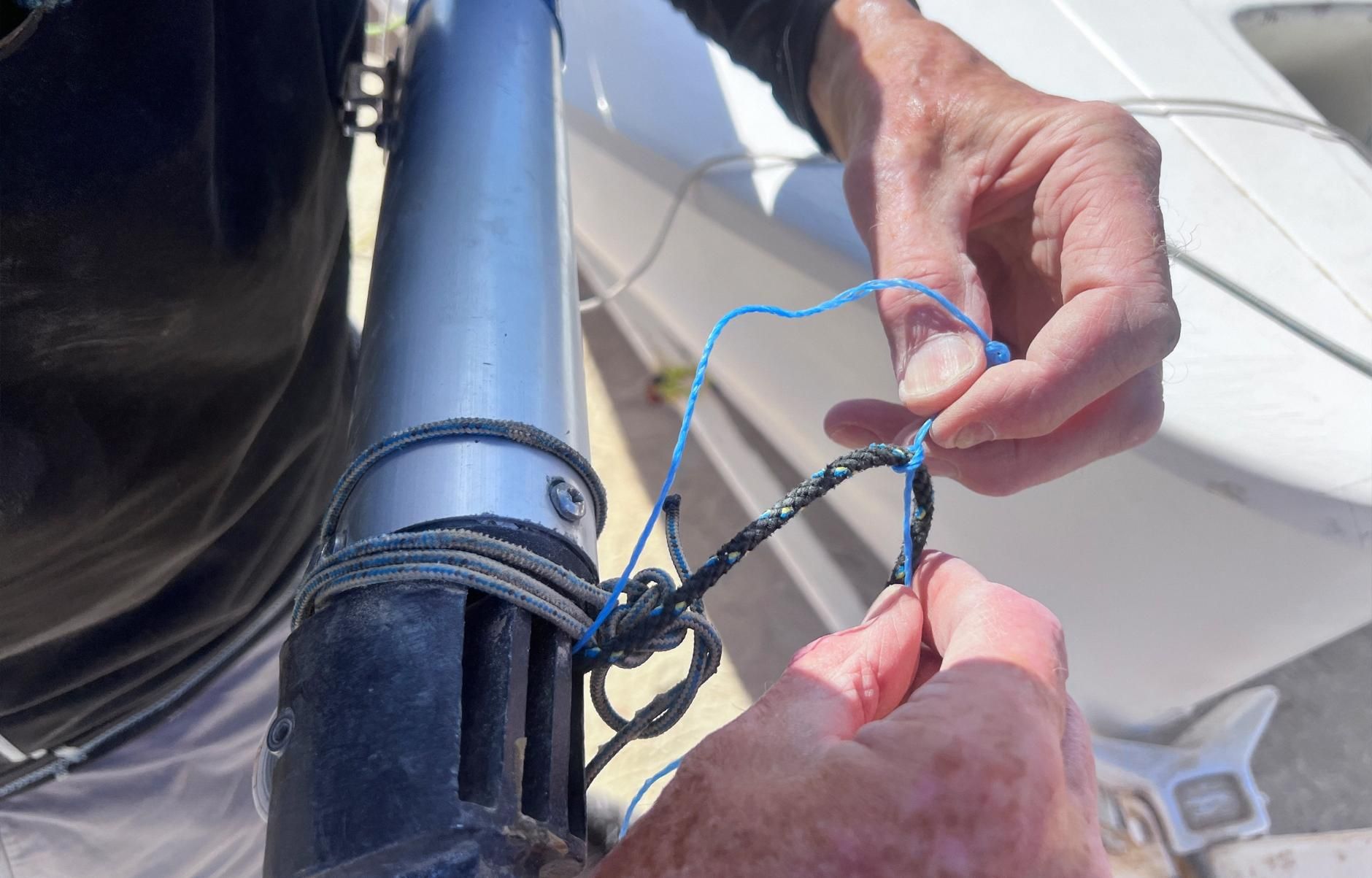Person's hands tying a blue rope around a black, cylindrical object, likely part of a boat mast.