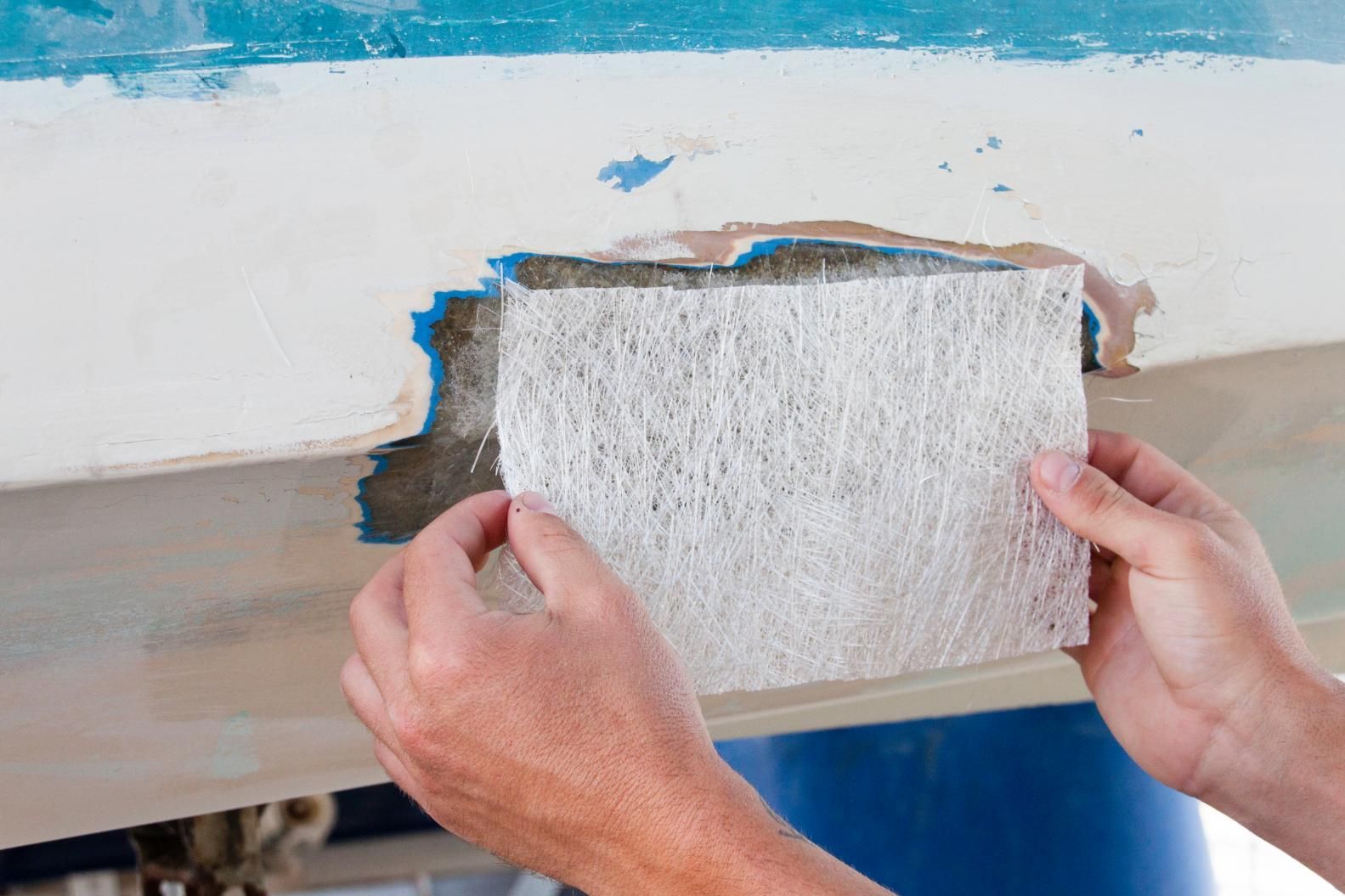 Person holding fiberglass cloth over a damaged area on a white and blue boat hull.