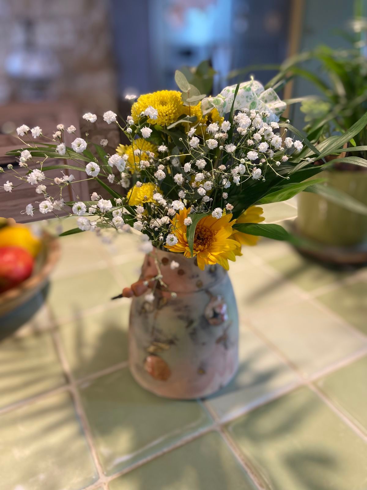 Des fleurs jaunes et du gypsophile dans un vase décoratif sur une table carrelée, près d'une coupe de fruits.