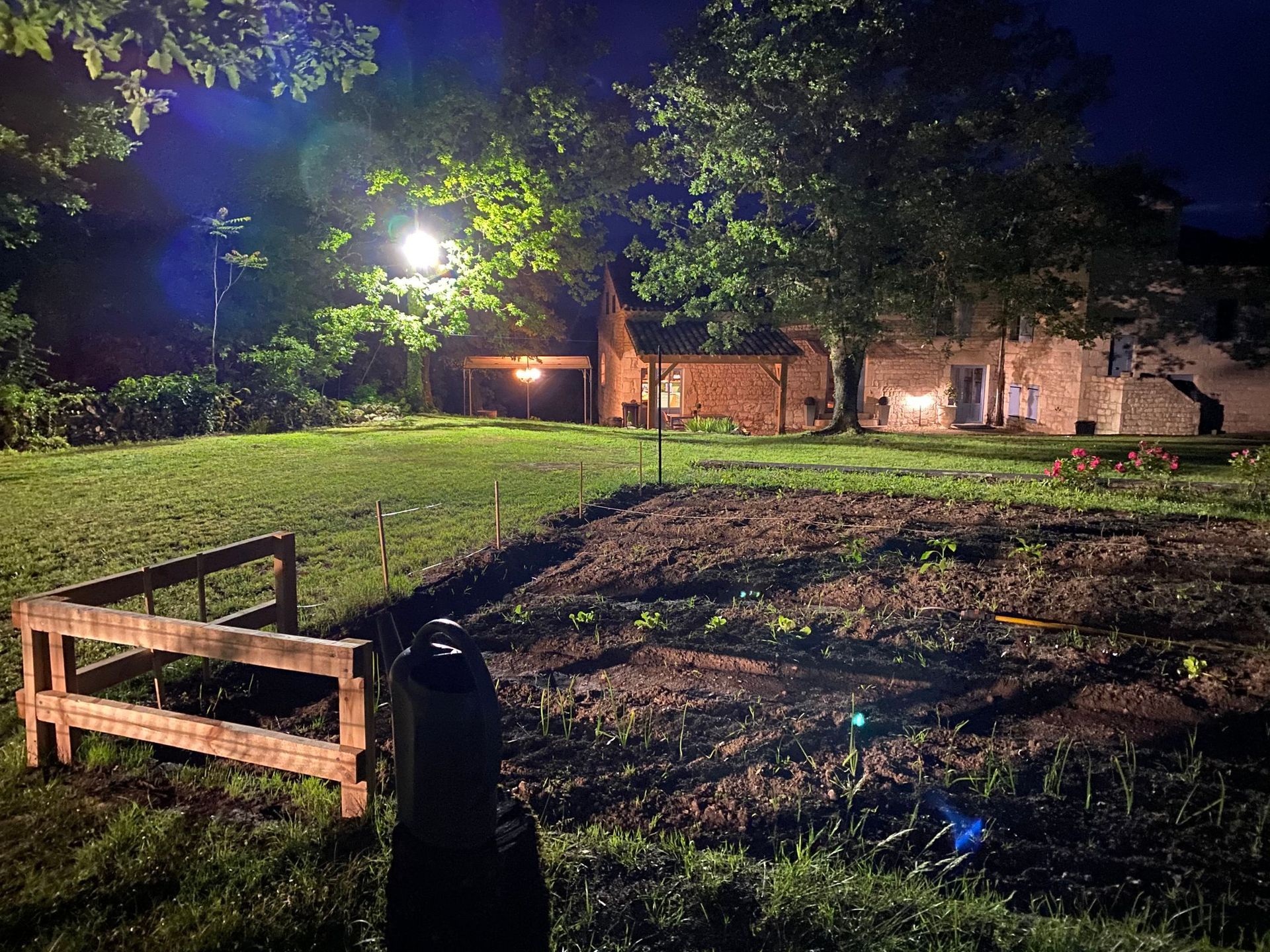 Une cour arrière la nuit, avec un jardin au premier plan, éclairé par les lampadaires et les lumières de la maison.