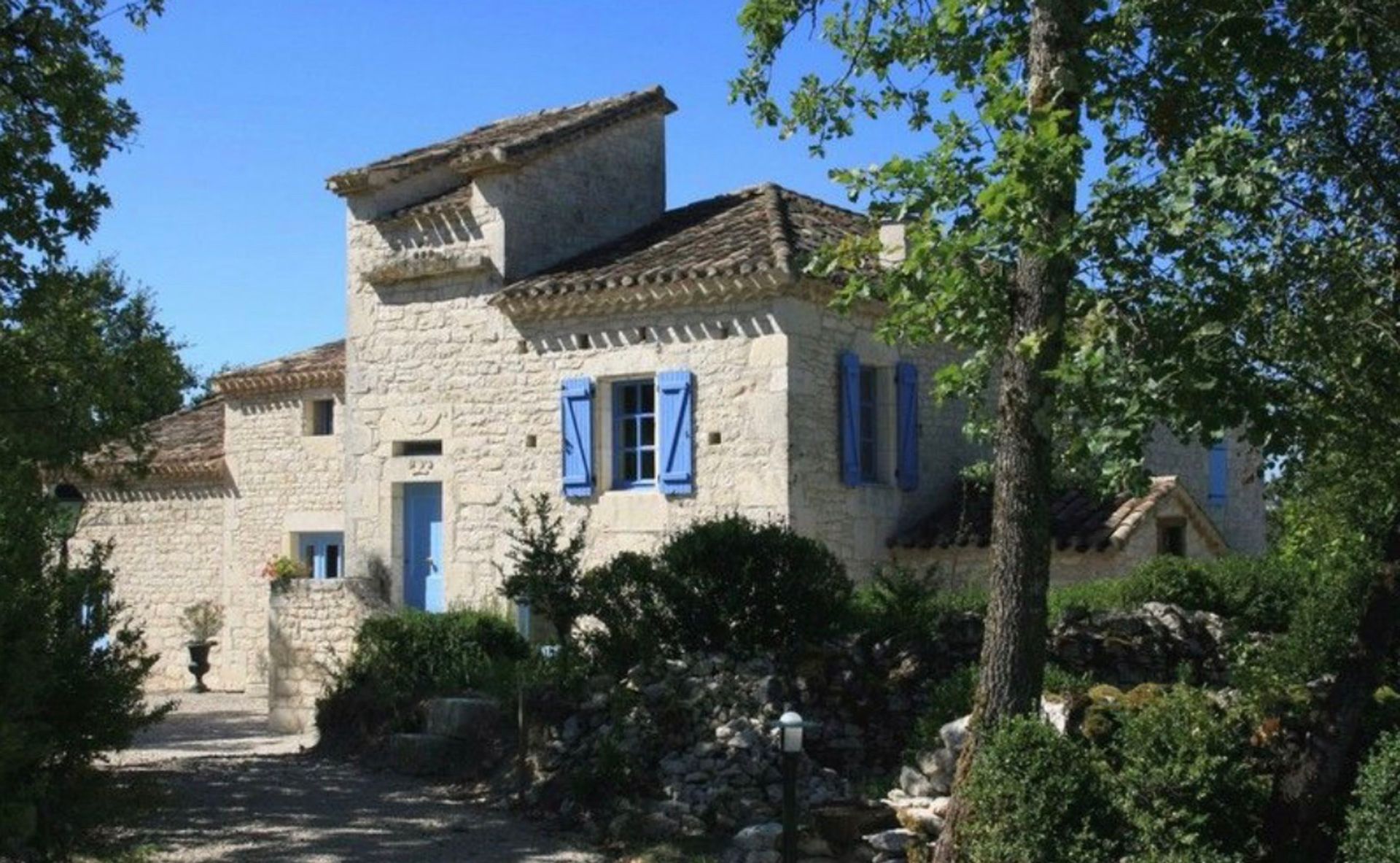 Maison en pierre aux volets bleus, entourée d'arbres sous un ciel d'un bleu limpide.