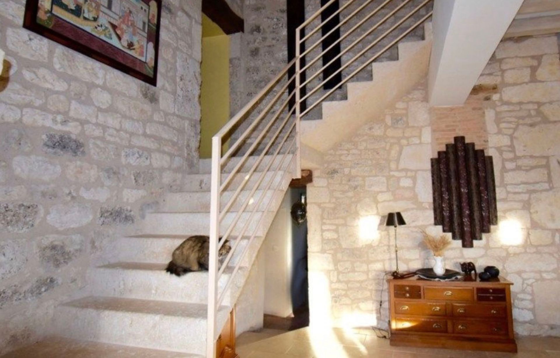 Intérieur aux murs de pierre avec un escalier et un chat se reposant sur une marche. Le mobilier et la décoration en bois apportent une touche d'élégance.