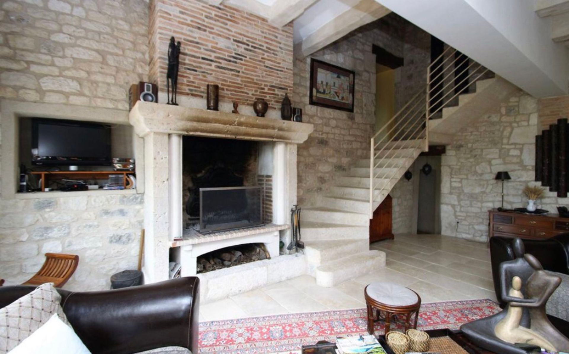 Salon rustique avec murs en pierre, cheminée, escalier et éléments décoratifs.