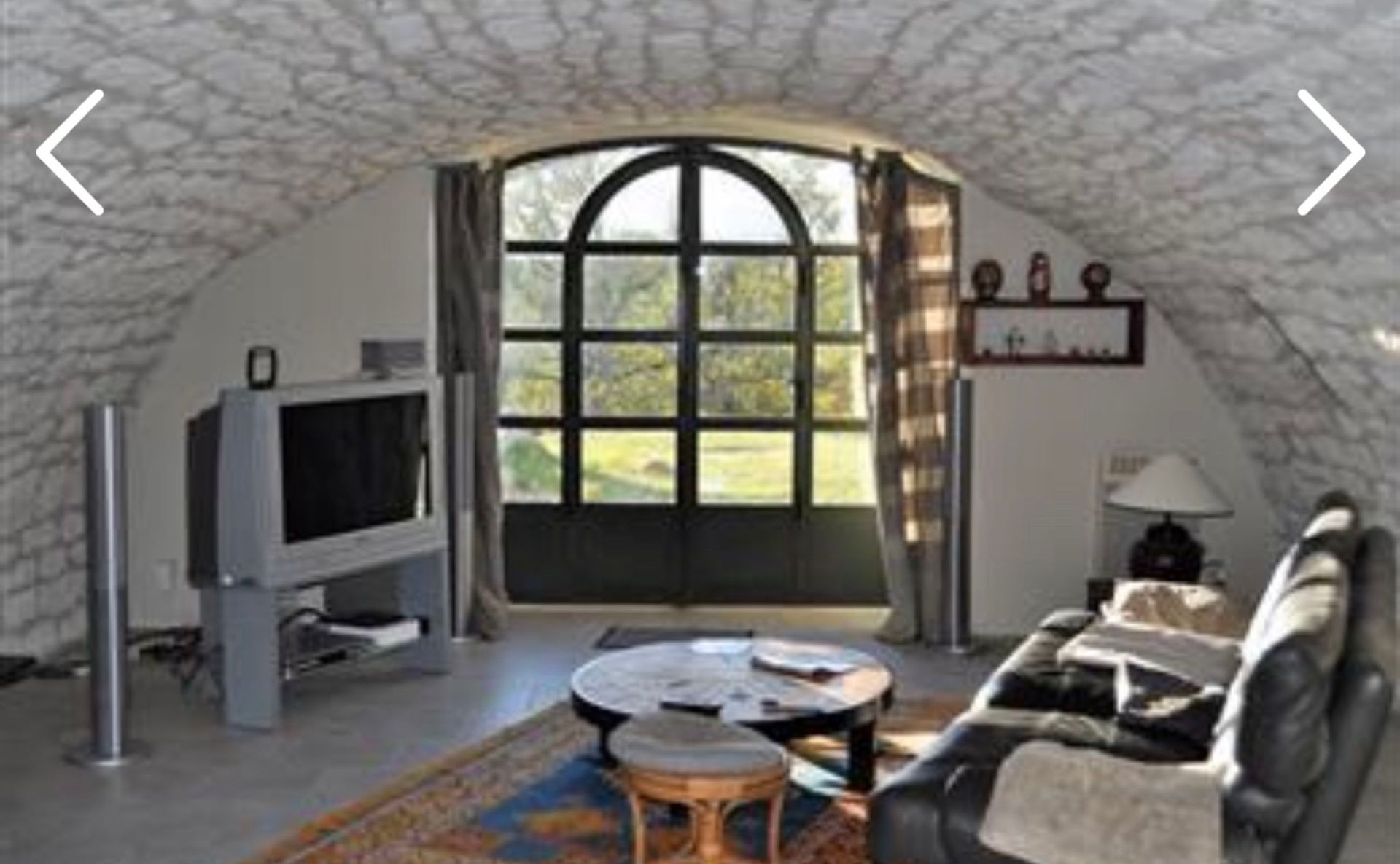 Salon avec plafond voûté et grande fenêtre, canapé en cuir noir, télévision et table basse ronde.