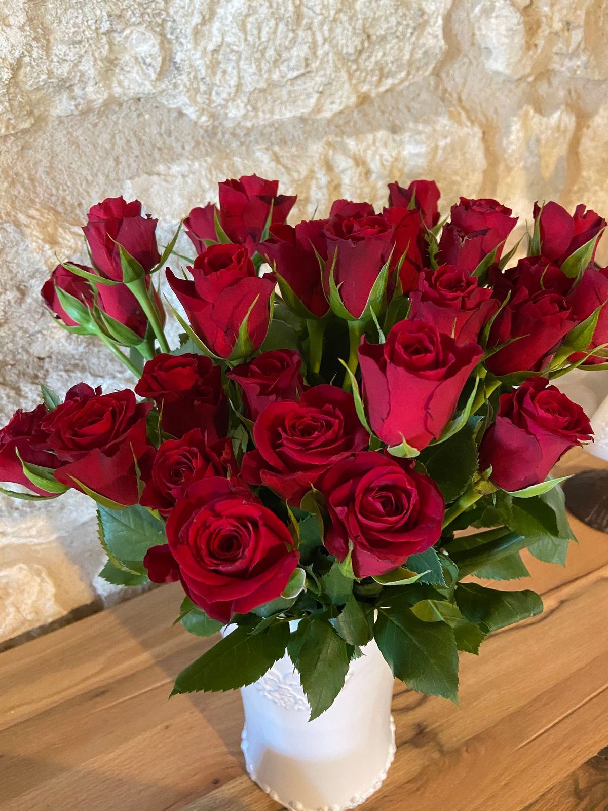 Des roses rouges dans un vase blanc, posées sur une surface en bois devant un mur de pierre.