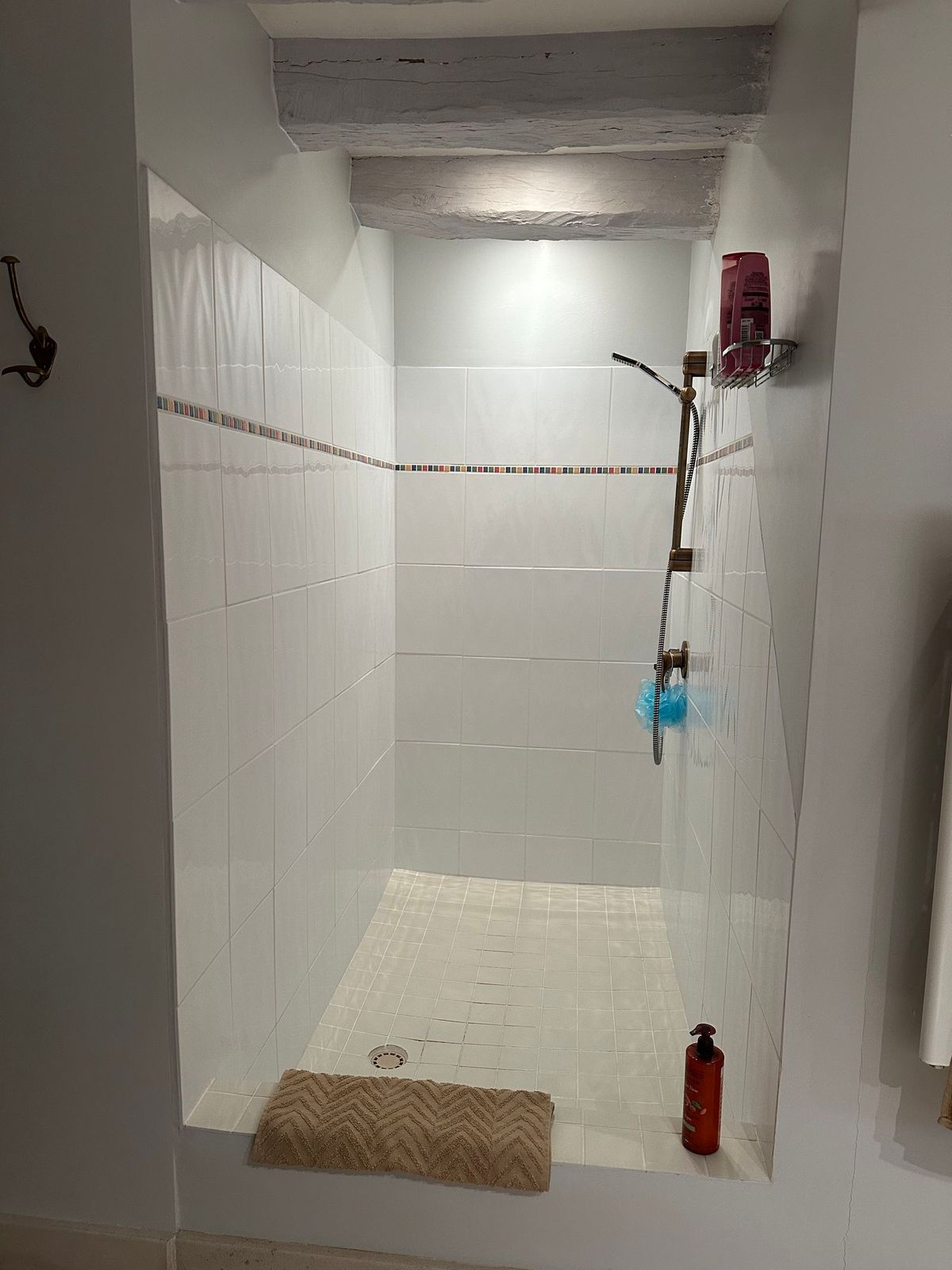 Douche carrelée de blanc avec pommeau effet pluie, flacons et tapis de bain. Poutres apparentes au plafond.