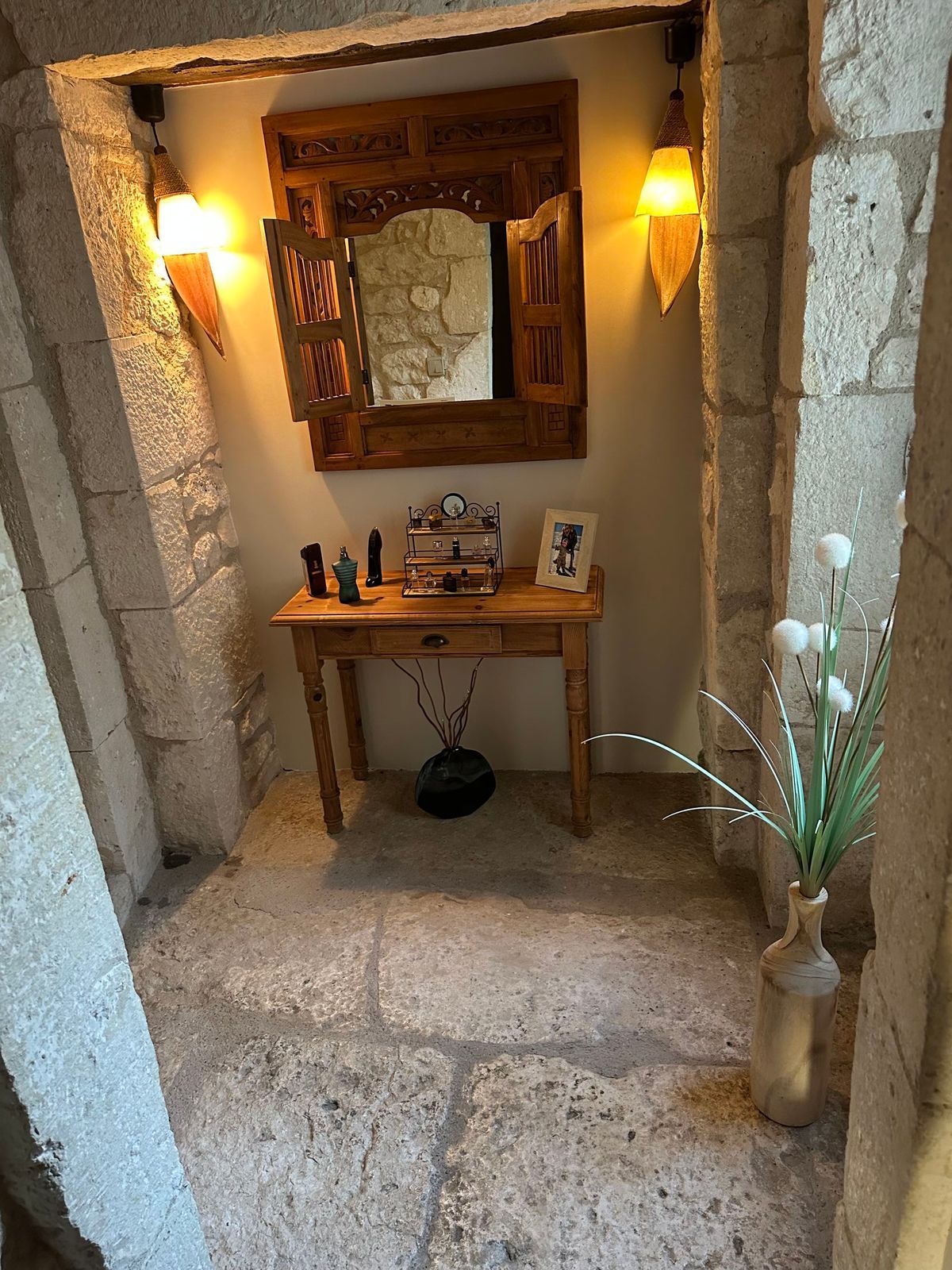 Étroite alcôve avec une table en bois, un miroir orné et des murs en pierre. Éclairée par des appliques et un vase de fleurs blanches.
