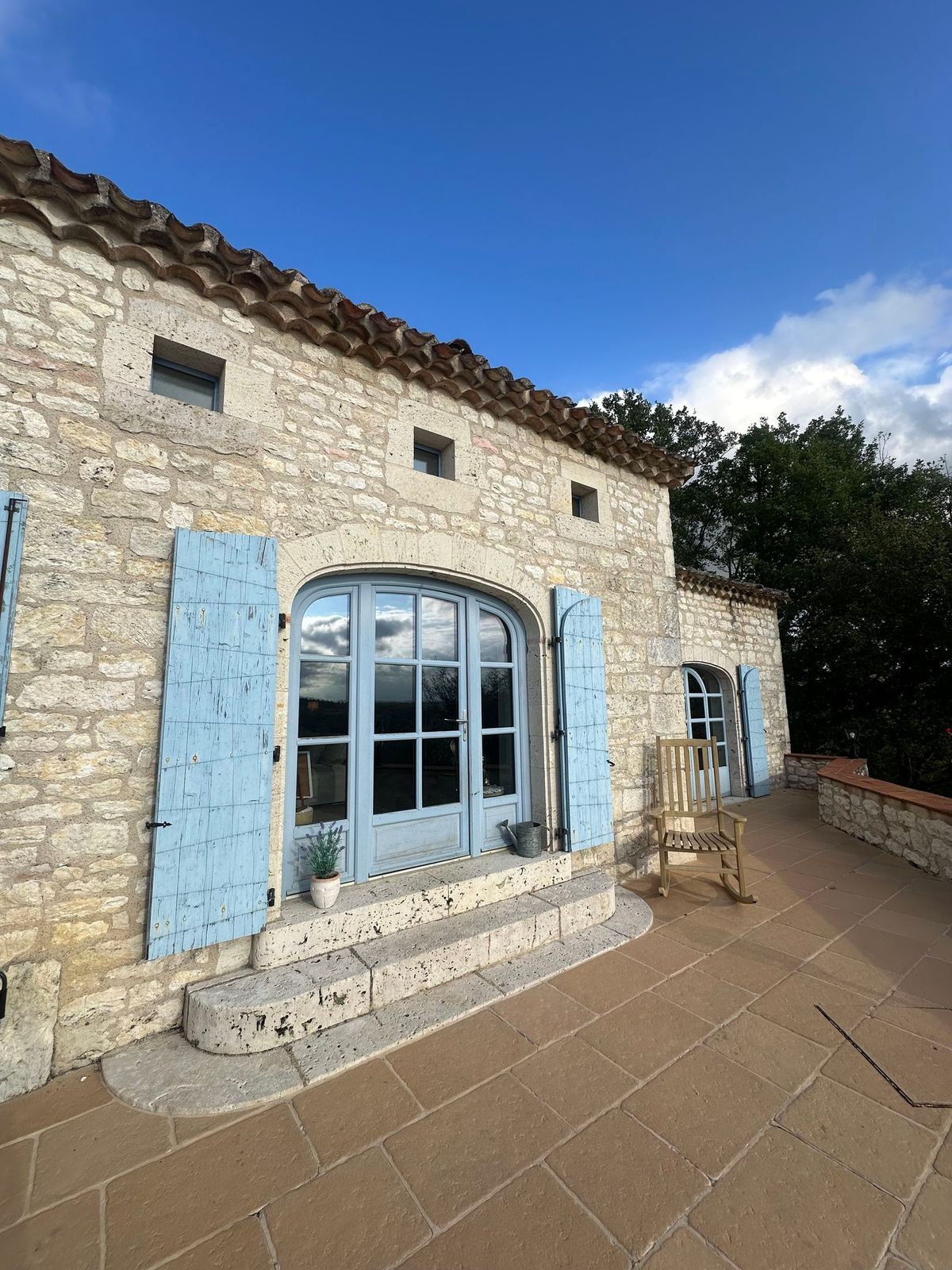 Maison en pierre avec volets bleus et fenêtre cintrée sur une terrasse ensoleillée.
