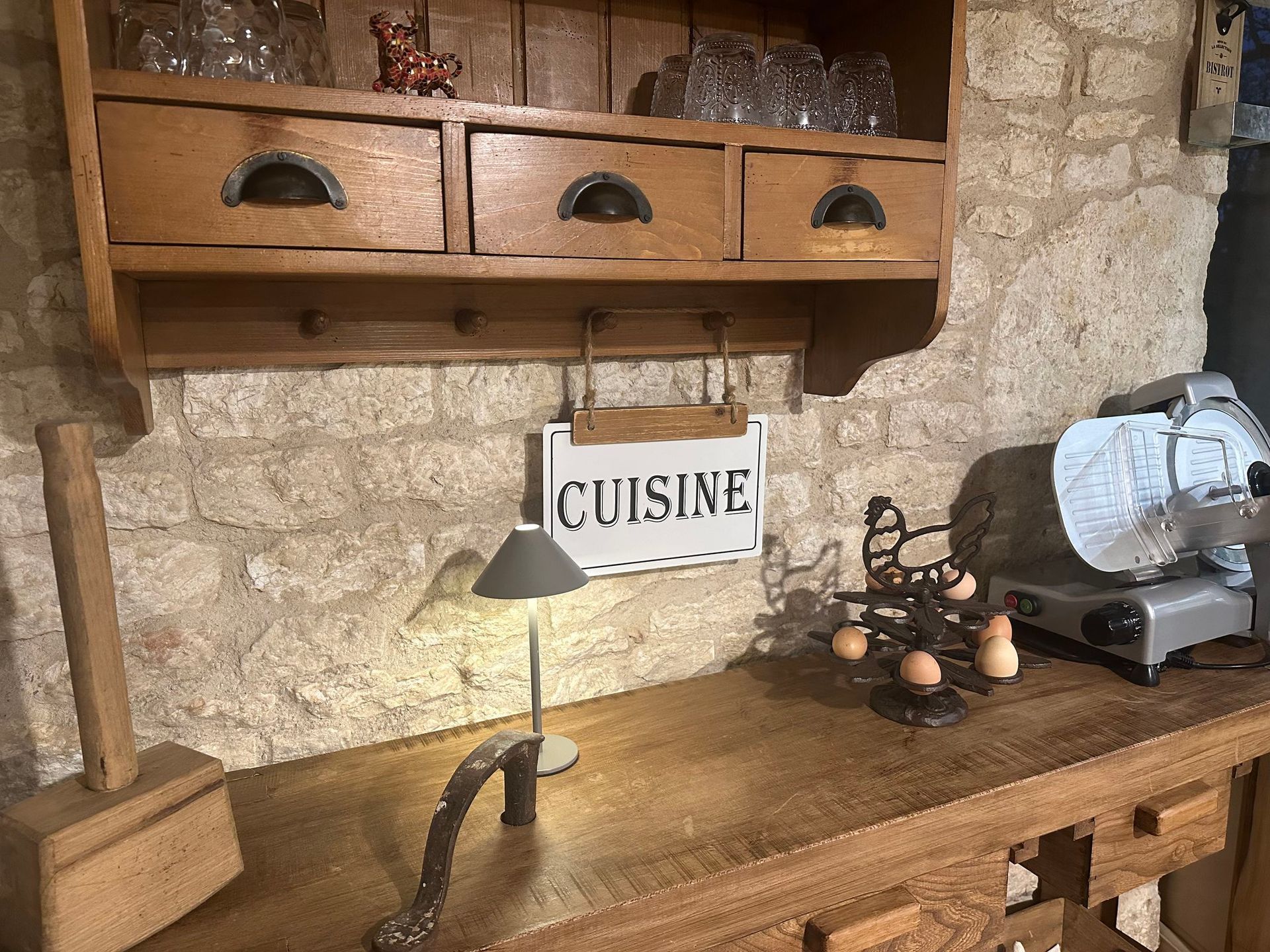 Plan de travail de cuisine avec étagères en bois, tiroirs et panneau « CUISINE ». Comprend une lampe, des œufs et une trancheuse.