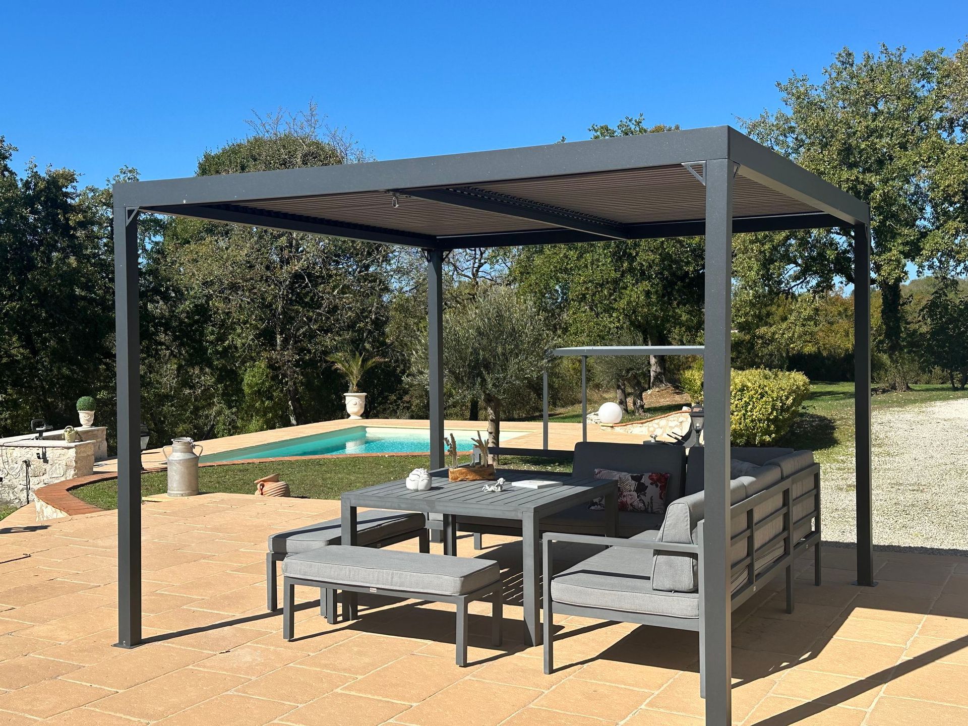 Pergola grise au-dessus d'un coin salon extérieur avec une piscine en arrière-plan par une journée ensoleillée.