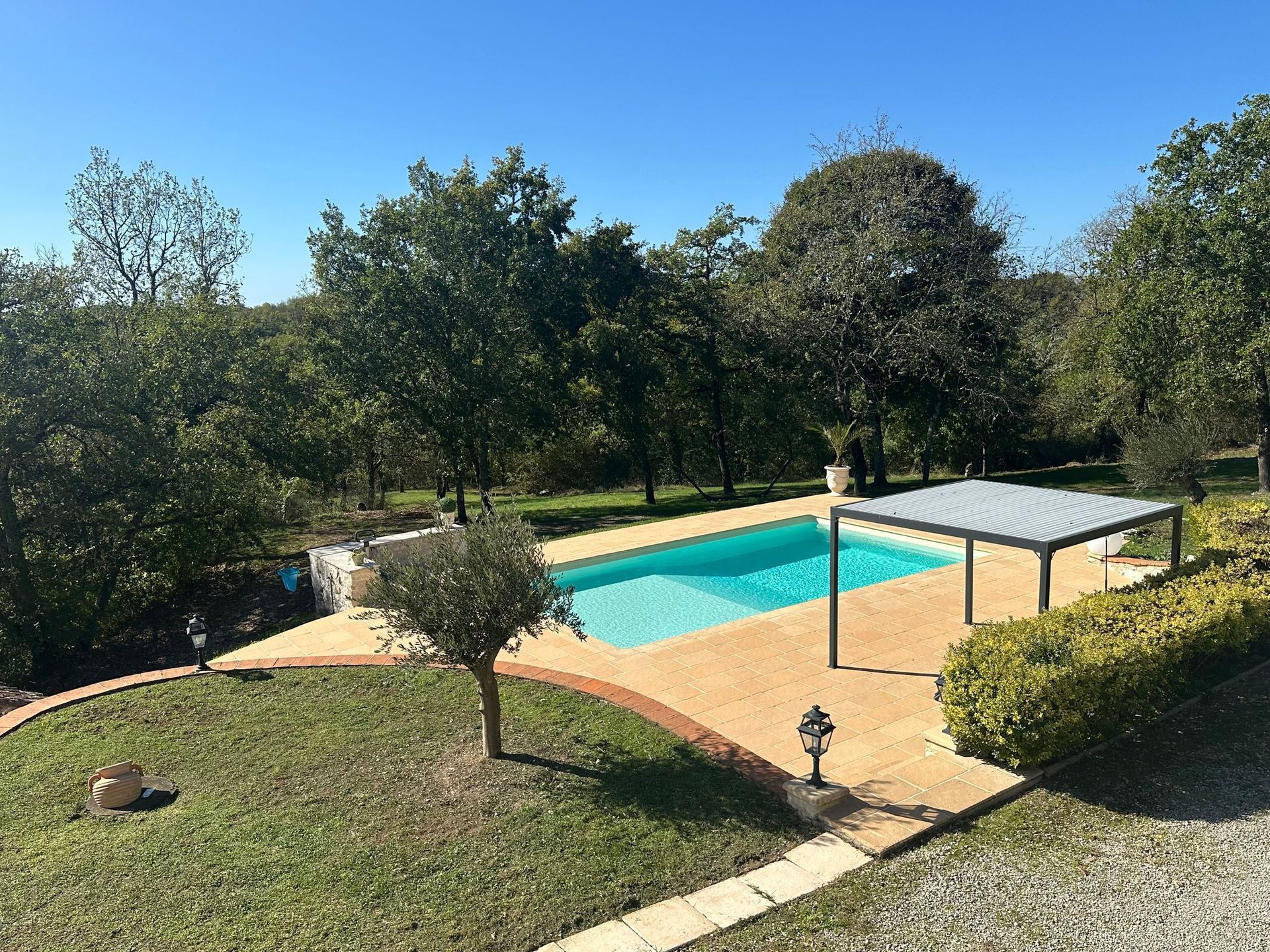 Une piscine rectangulaire dans un jardin ensoleillé, entourée de pierres beiges et d'arbres luxuriants. Un auvent métallique offre de l'ombre.