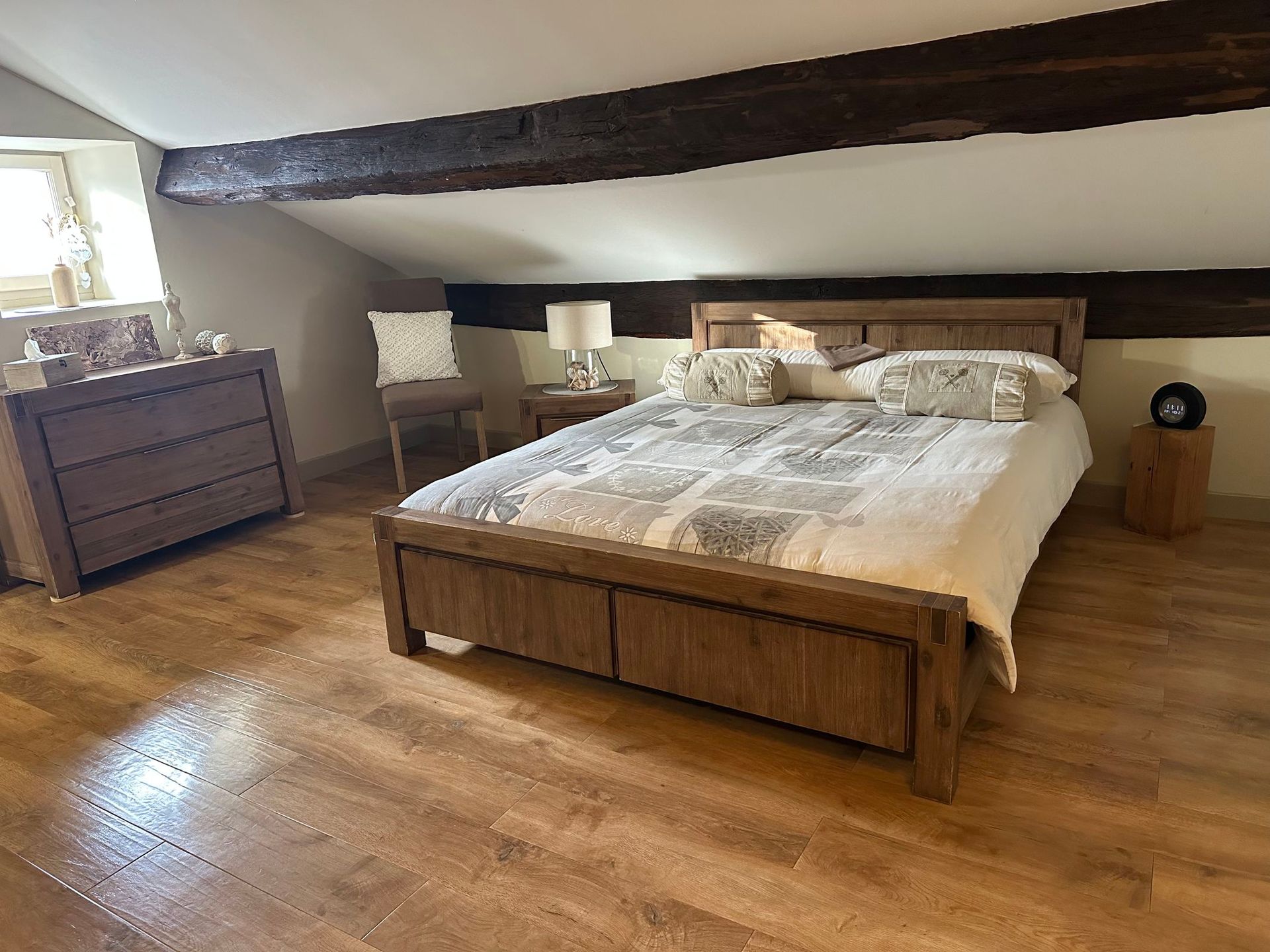 Chambre en bois avec lit, commode, chaise, table de chevet ; plafond mansardé avec poutre apparente ; parquet.