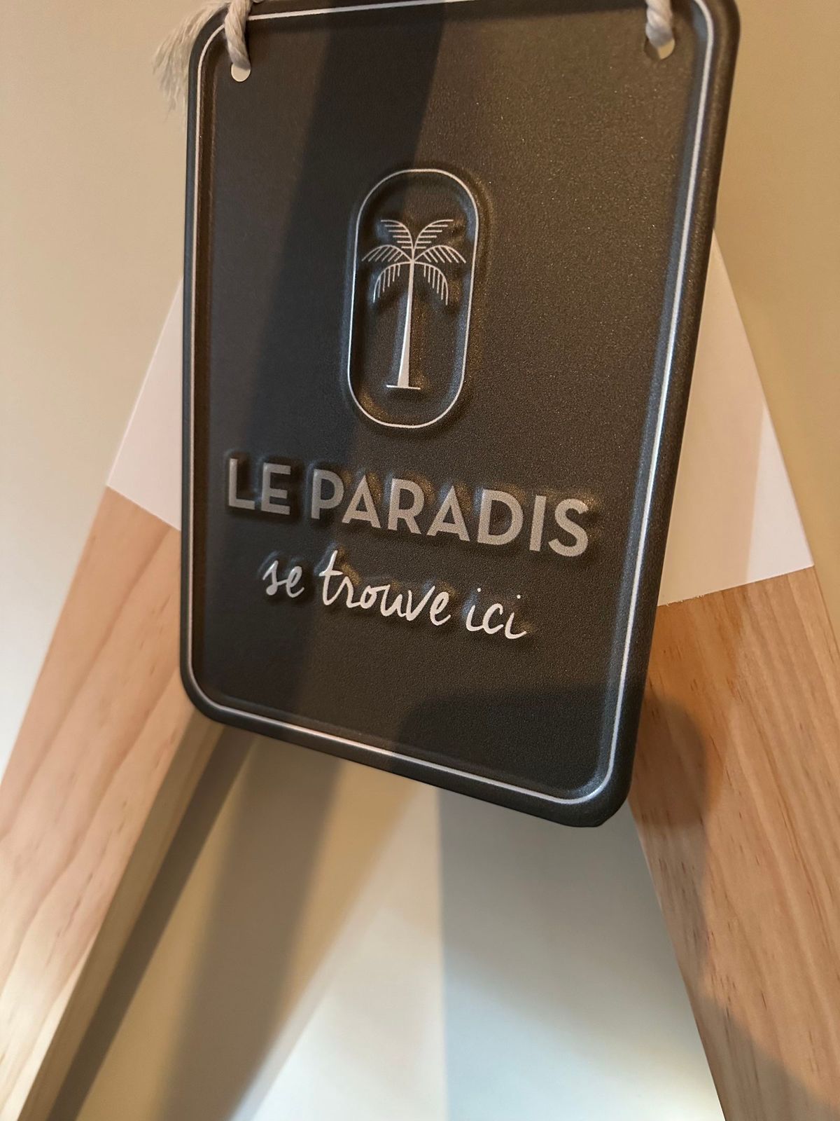 Panneau noir avec l'inscription « LE PARADIS se trouve ici » en lettres argentées et une icône de palmier, accroché sur un cadre en bois.