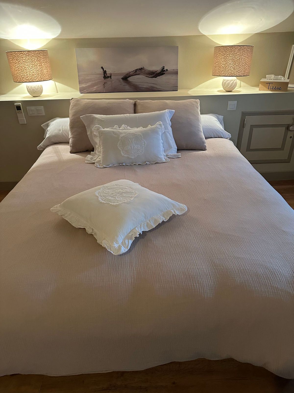 Chambre avec lit recouvert de draps rose pâle et d'oreillers blancs. Deux lampes encadrent un tableau accroché au mur.