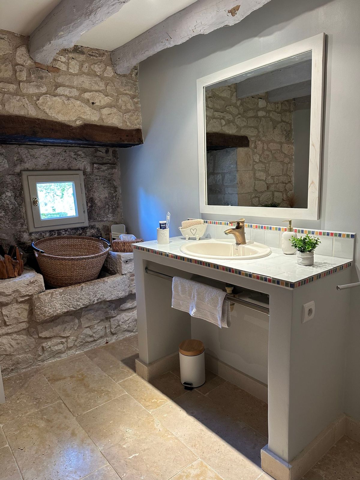 Salle de bains avec murs en pierre, un grand miroir, un lavabo blanc et une fenêtre.