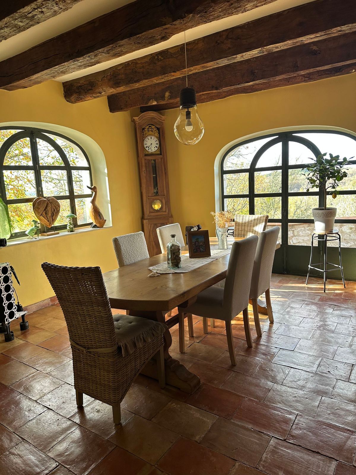 Salle à manger avec table en bois, chaises, fenêtres cintrées, horloge grand-père et poutres apparentes.