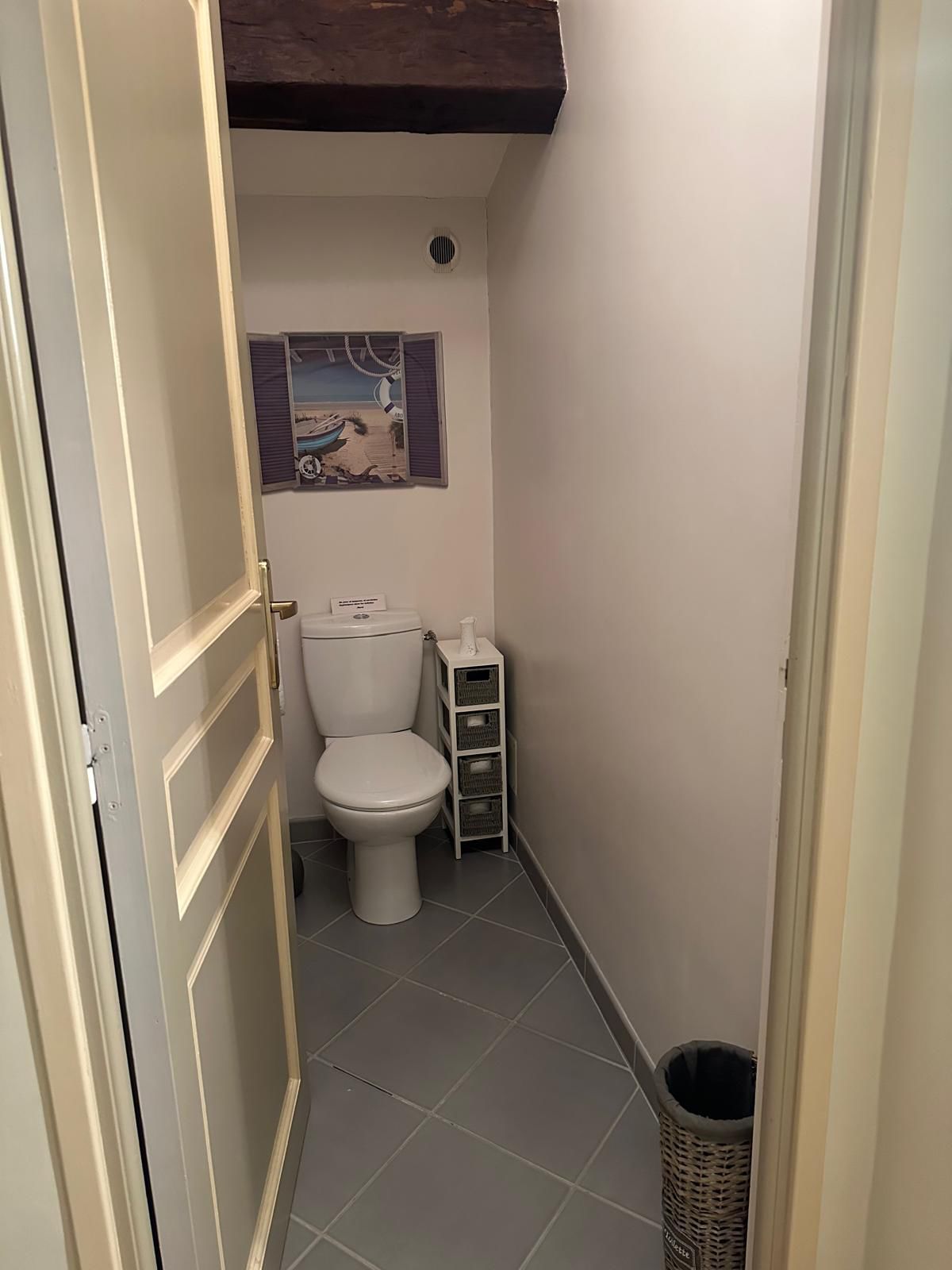 Toilettes dans une petite salle de bains. Murs blancs, sol carrelé gris, cadre de porte ouvert, petit meuble de rangement, poubelle.