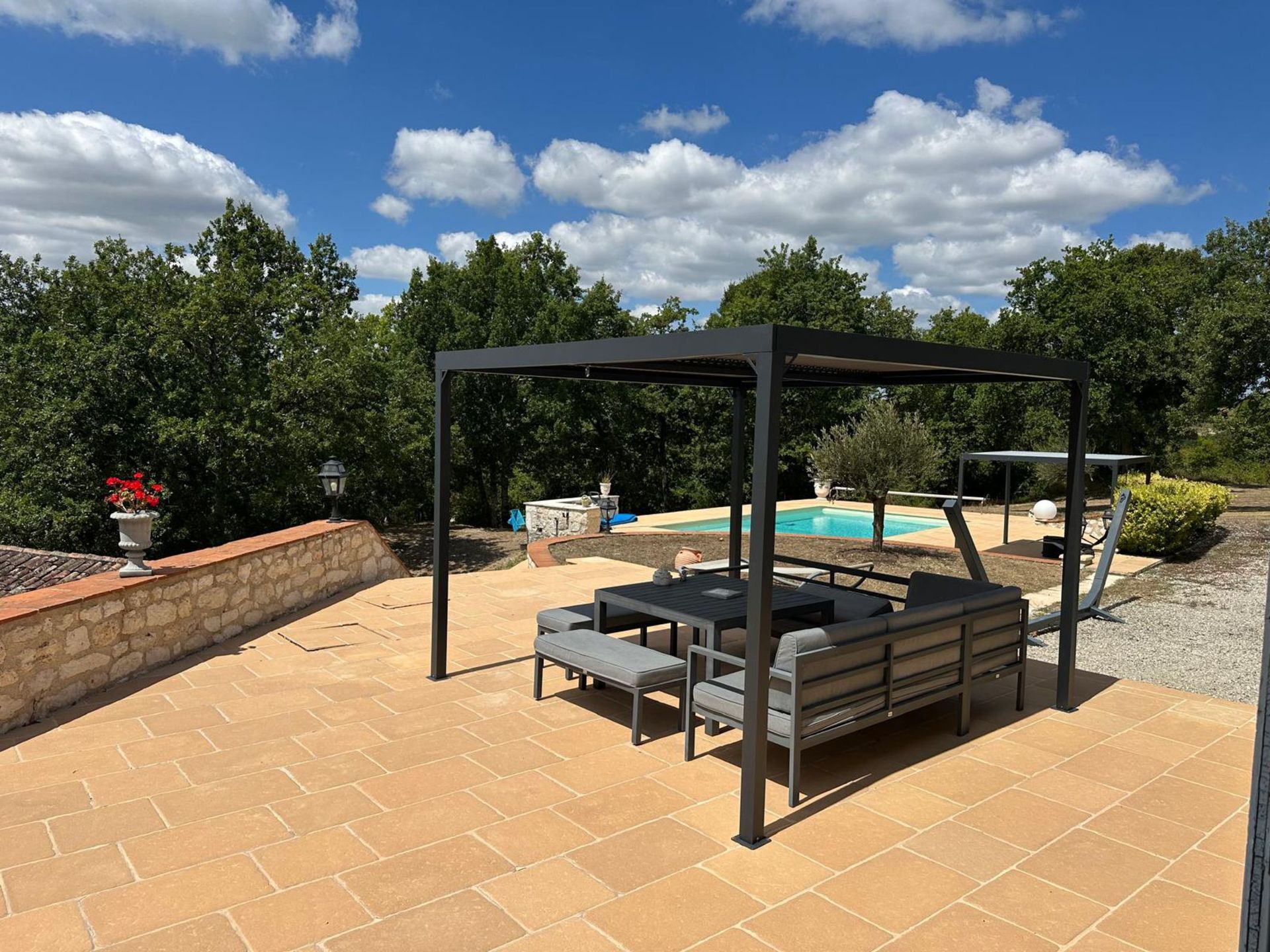 Terrasse avec mobilier de jardin moderne sous une pergola, arbres, piscine et ciel bleu.