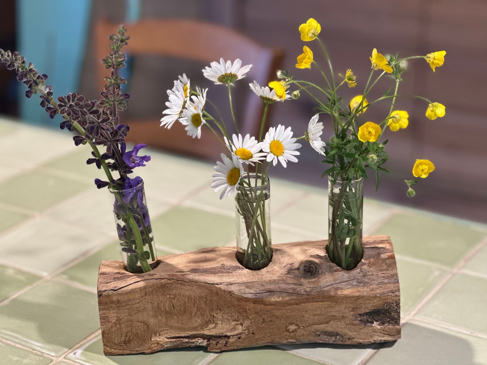 Trois petits vases remplis de fleurs sauvages, disposés dans un support en bois rustique. Des marguerites, des fleurs jaunes et des fleurs violettes.