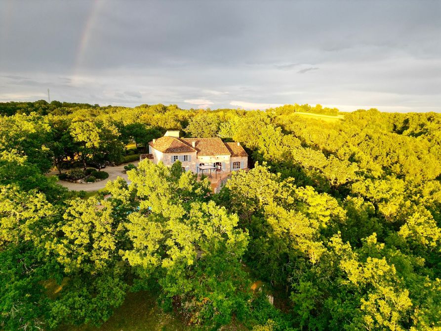 Une maison en pierre nichée dans une forêt dense et verdoyante, partiellement dissimulée par les arbres, avec un arc-en-ciel qui se dessine au-dessus.