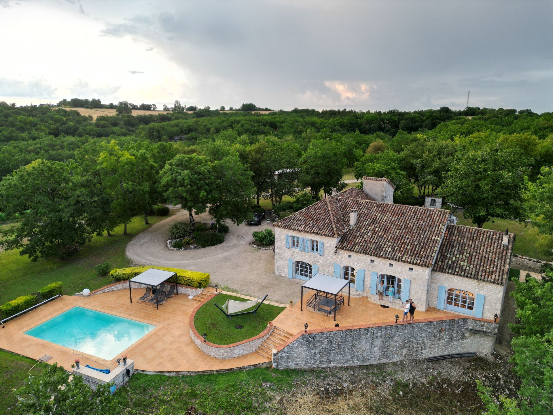 Maison en pierre avec piscine dans un cadre verdoyant luxuriant sous un ciel nuageux.