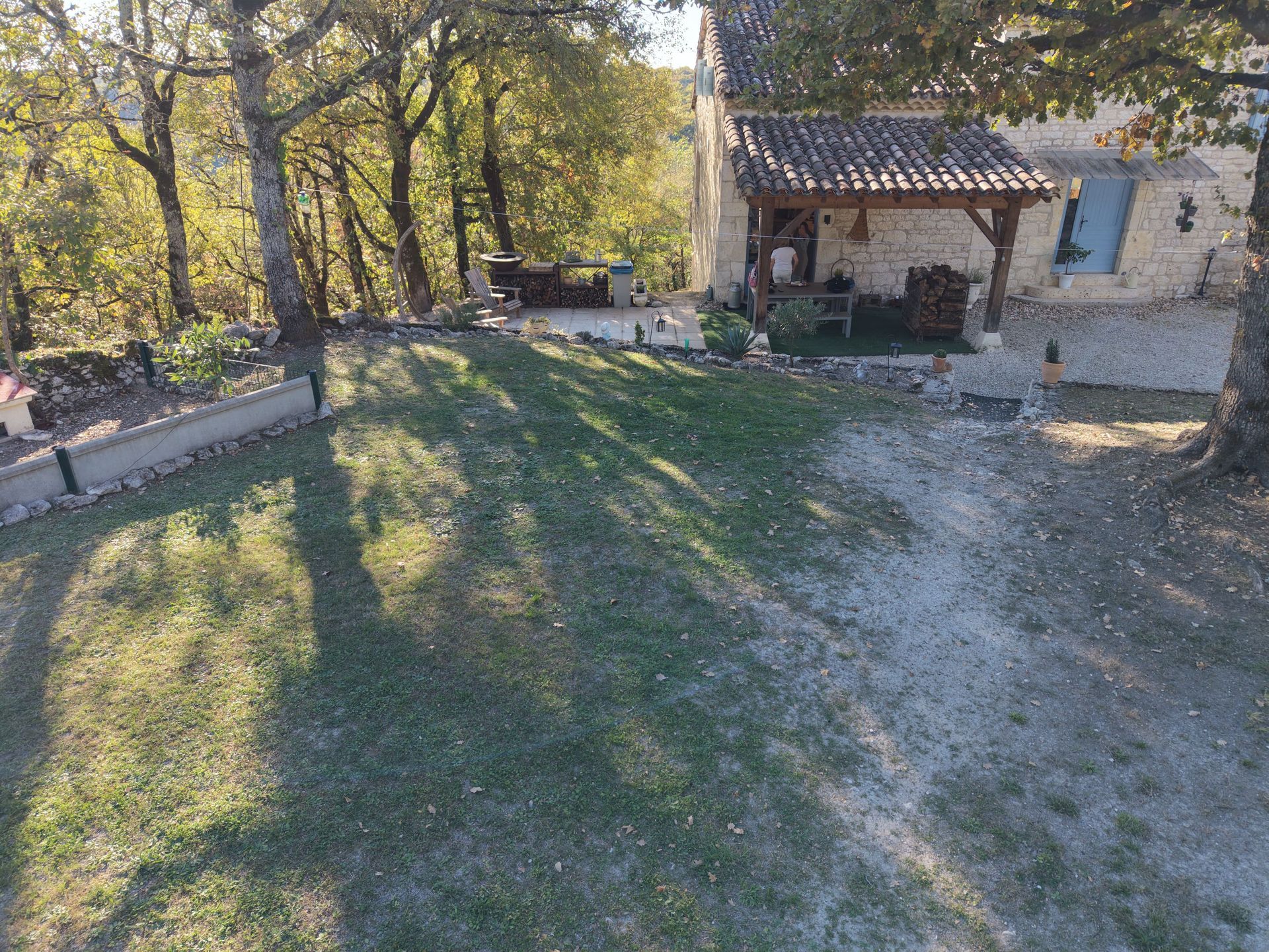 Maison en pierre avec toit de tuiles et porche en bois, située dans une cour avec pelouse et arbres.