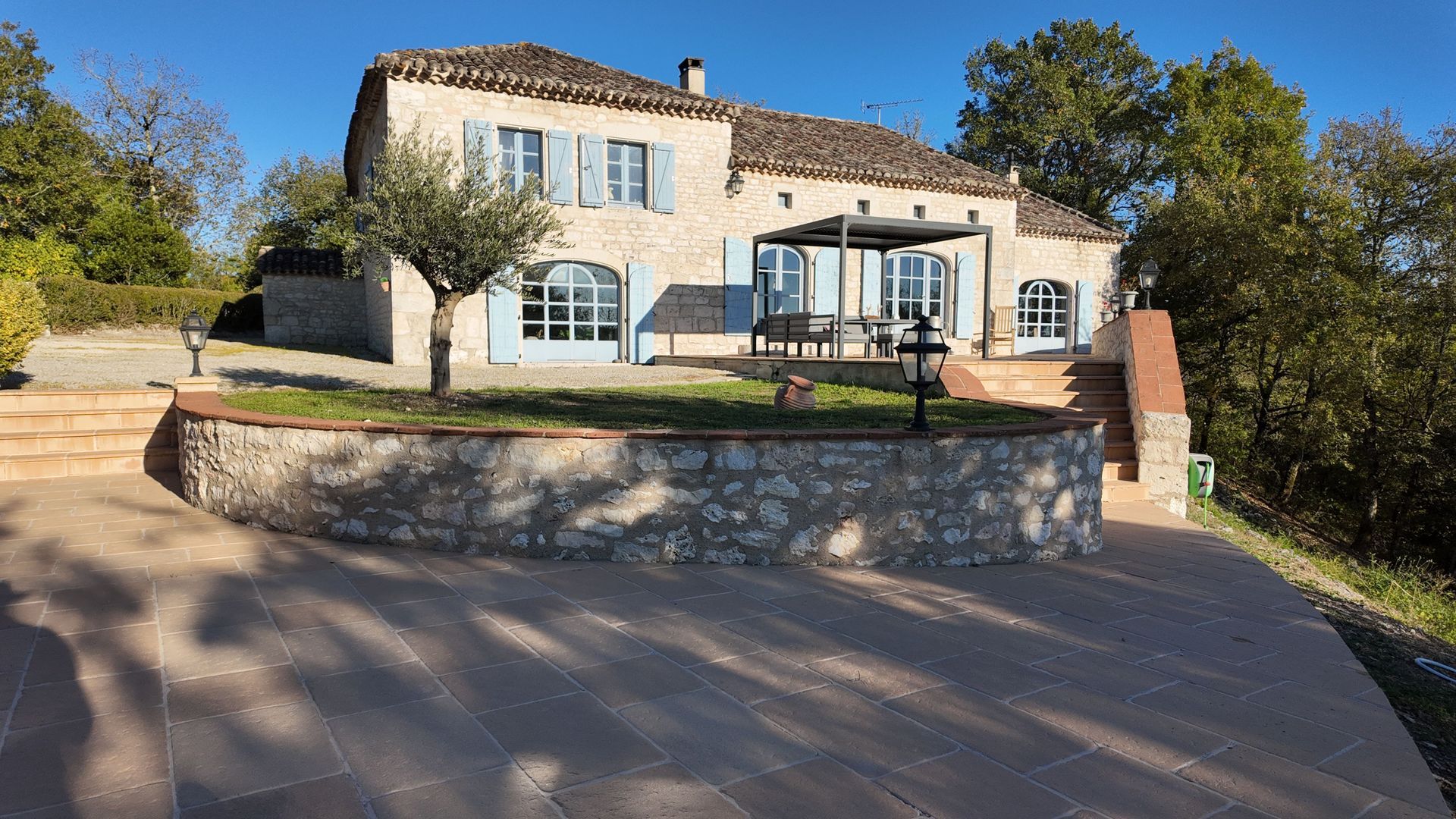 Maison en pierre aux volets bleus, située sur une colline, avec une terrasse et un petit jardin.