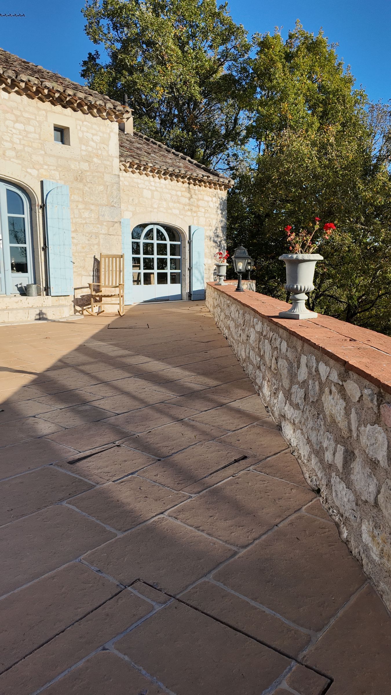 Terrasse en pierre avec un muret, bâtiment aux fenêtres cintrées et arbre en arrière-plan.