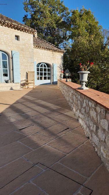 Maison en pierre aux volets bleus, avec une terrasse et un mur de briques. Un vase de fleurs rouges est posé sur le mur.