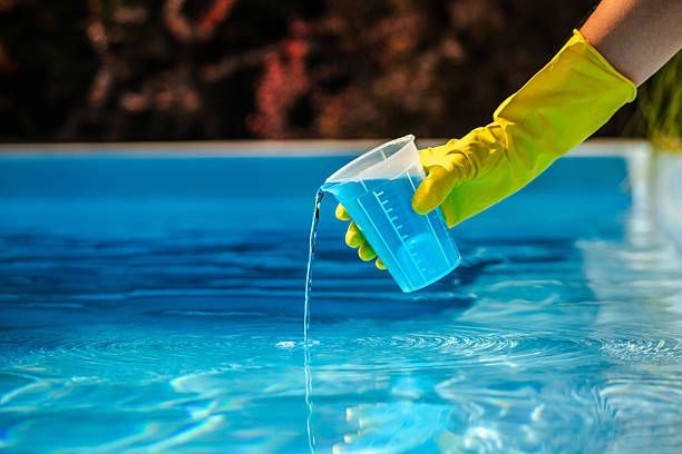 Gloved hand pouring blue pool water from a test cup into a swimming pool