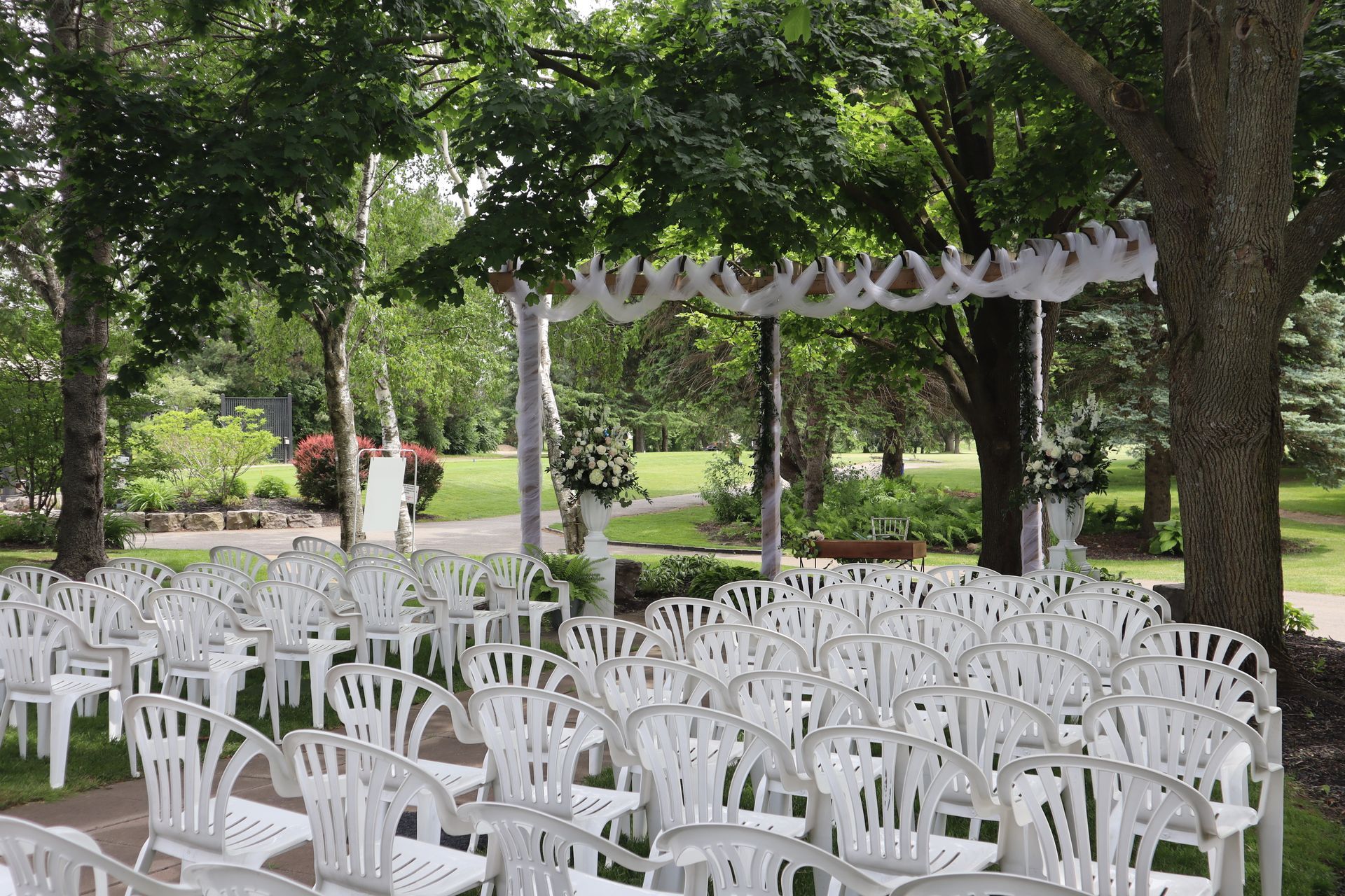 A row of white plastic chairs are lined up for an outdoor ceremony with an arbour with white drapery