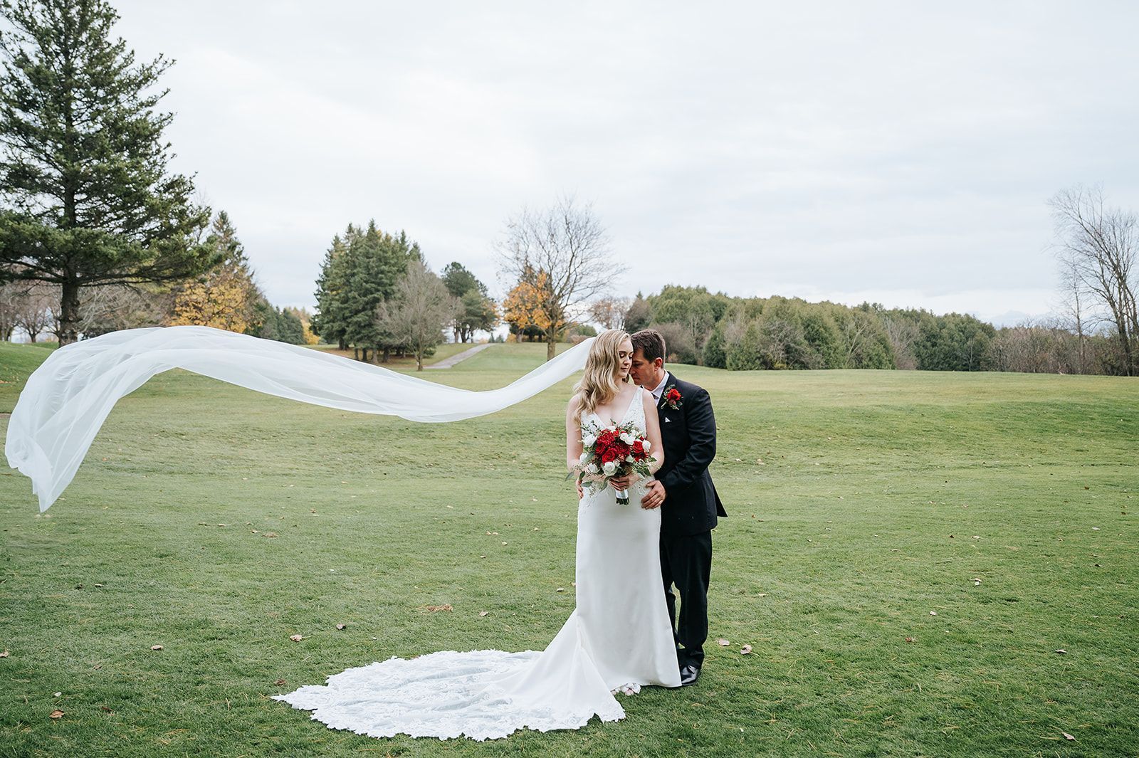 A bride and groom are standing in a field with bride's veil blowing in the wind.