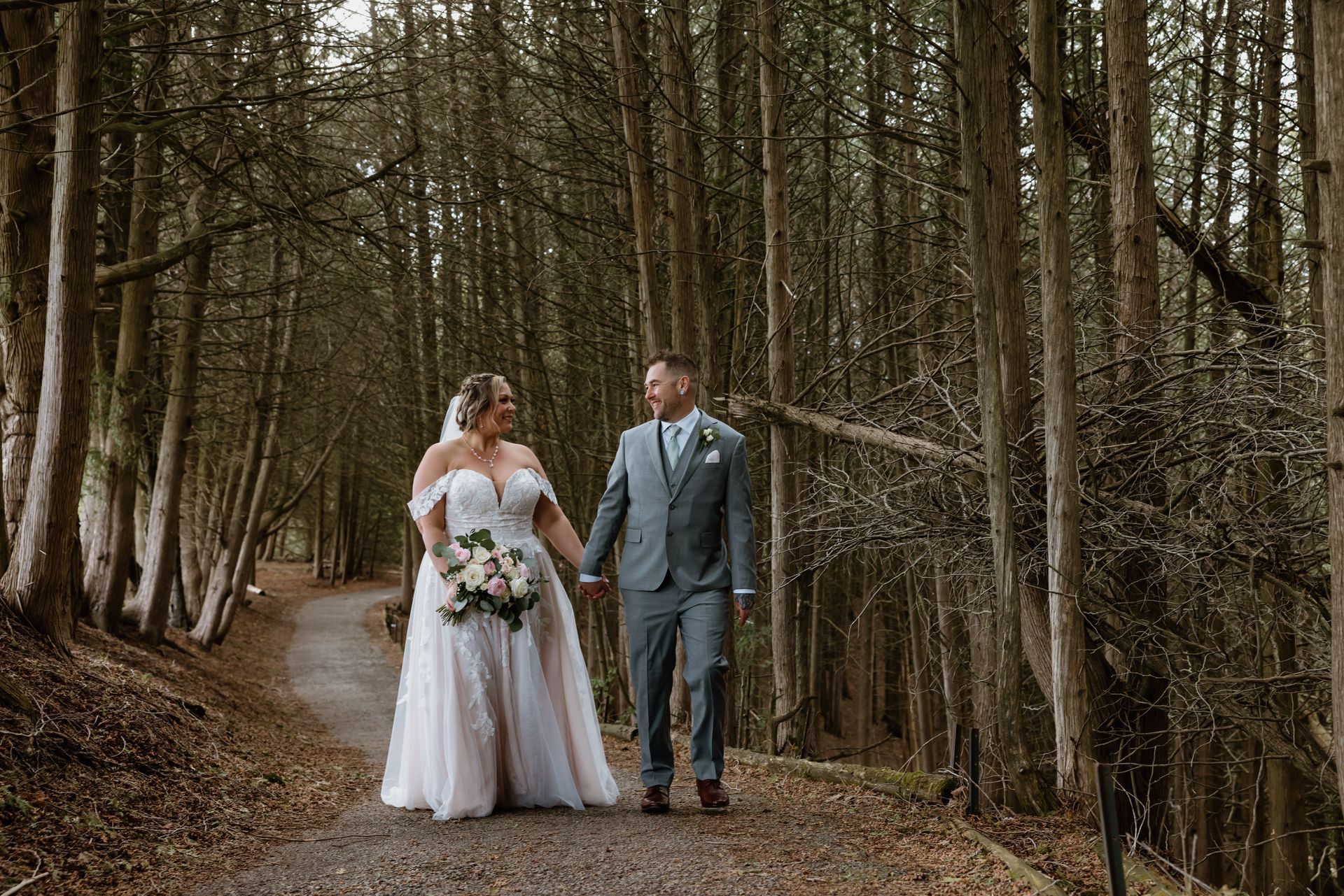 A bride and groom are walking down a path in the woods holding hands.