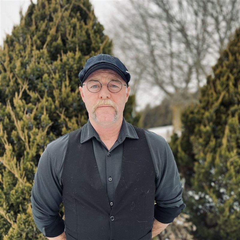 A man, Mike Ward, in a vest and hat is standing in front of a pine tree.