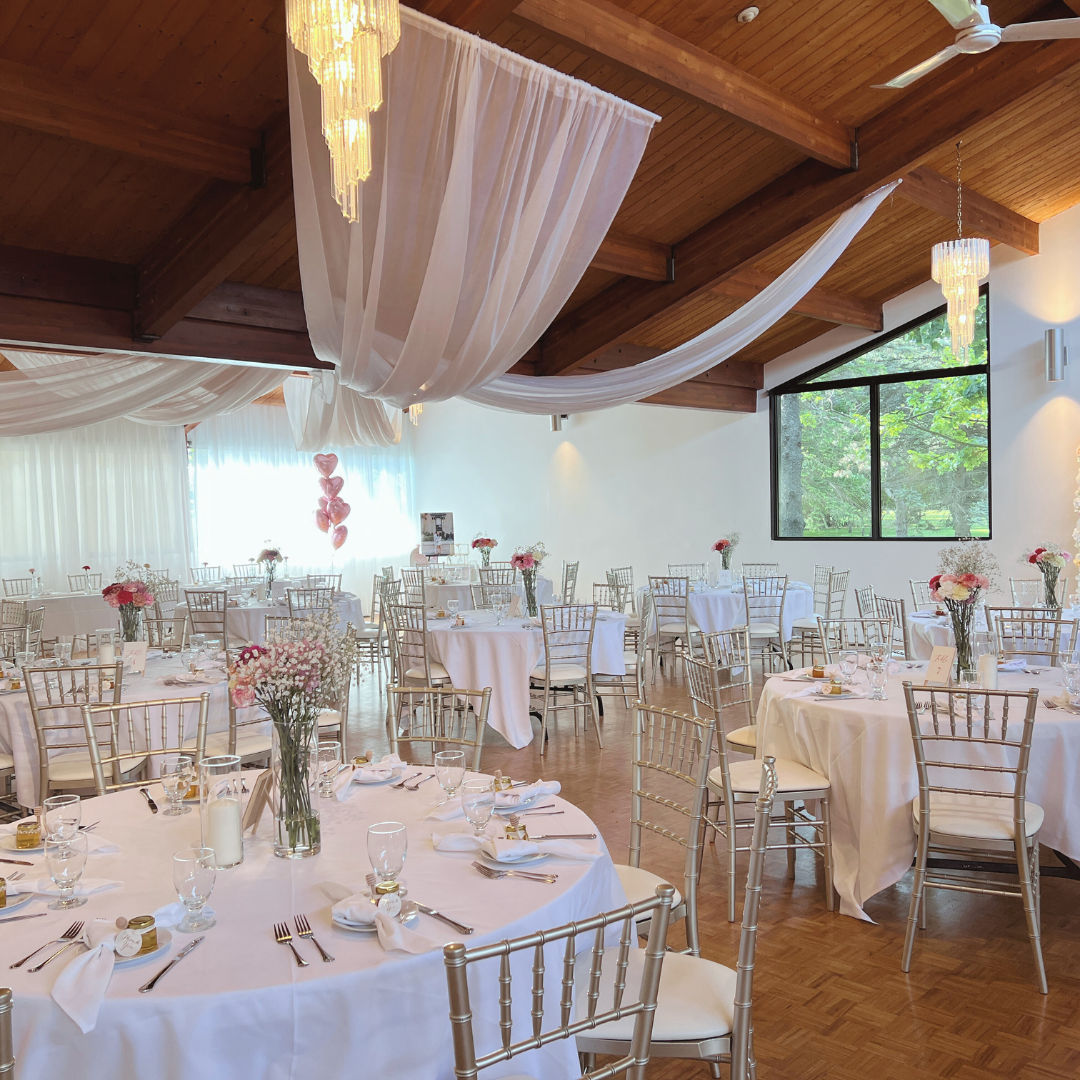 A large room with white tables and champagne chivari chairs with floral centerpieces setup for an event.