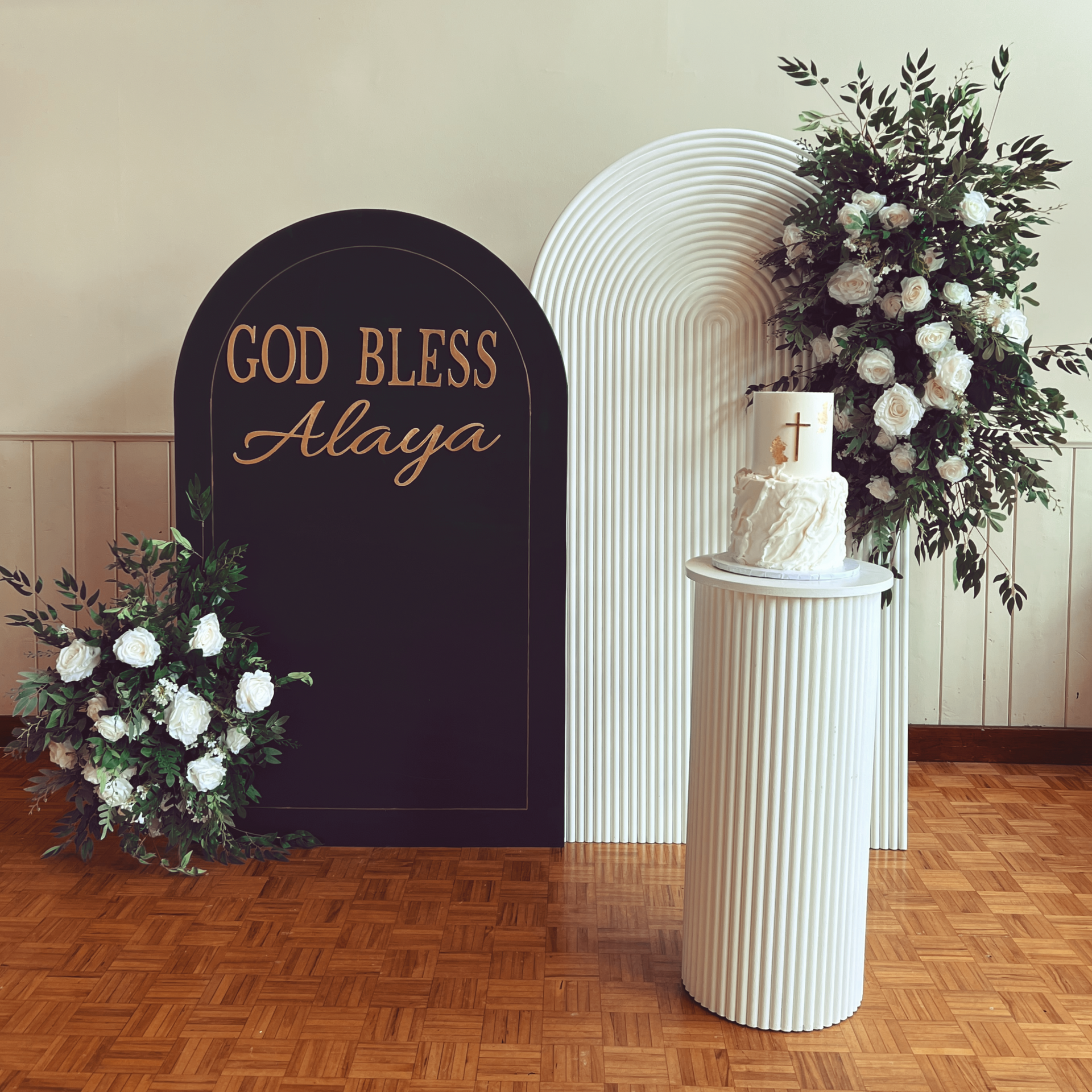 A cake on a white stand with a green arch reading 'God Bless Alaya' and a white floral arch