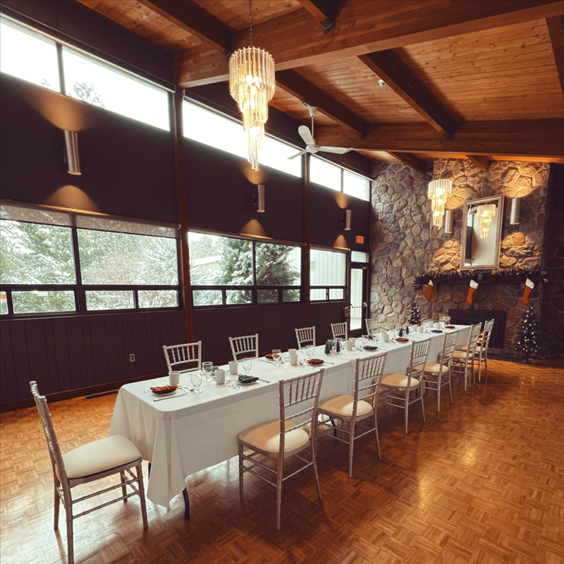 A large room with a long table and chairs in front of a stone fireplace and tall wood beam ceiling