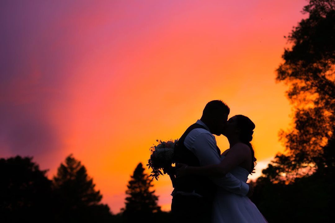 A bride and groom are kissing in front of a colorful sunset.