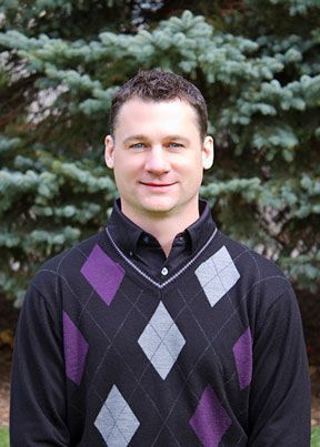 A man, Phil Brown, wearing a black and purple argyle sweater is standing in front of a pine tree.