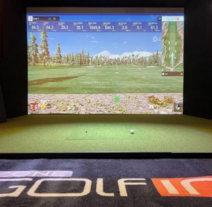 A close up of a golf simulator with a golf ball on the mat displaying a golf range on the screen.