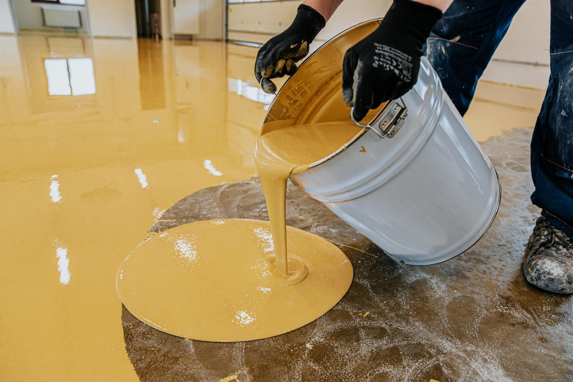 Person pouring yellow epoxy from a bucket onto a concrete floor, prepping for coating.