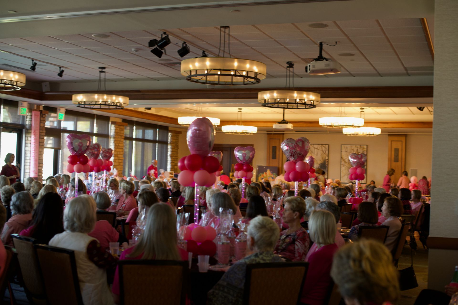 A large room filled with people sitting at tables with pink balloons on them