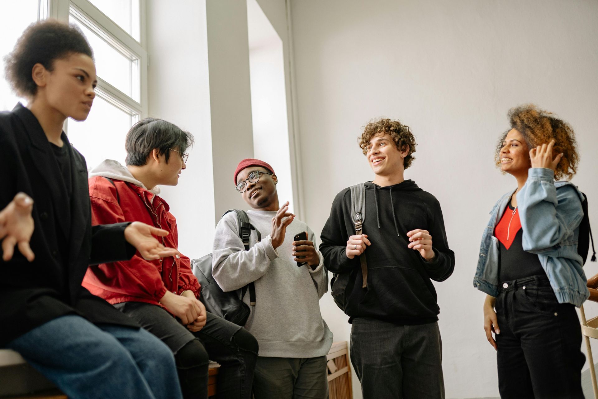 Five diverse students conversing in a well-lit room, two wearing backpacks.