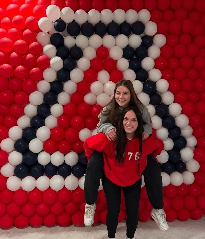 Two women are posing in front of a wall of red white and blue balloons