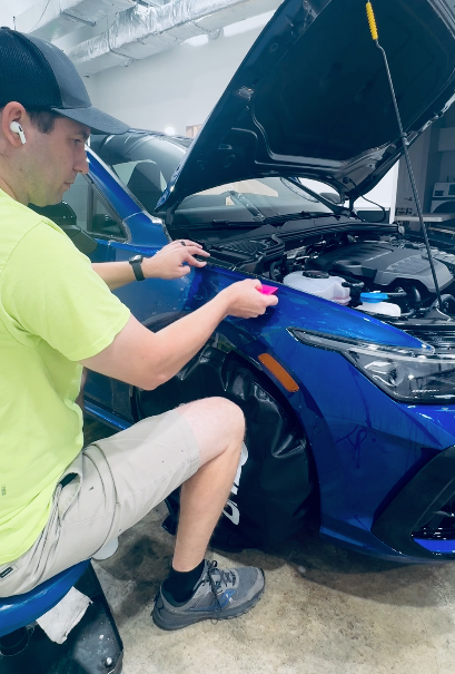 Man applying blue film to a blue car's fender, wearing a cap and kneeling inside a workshop.
