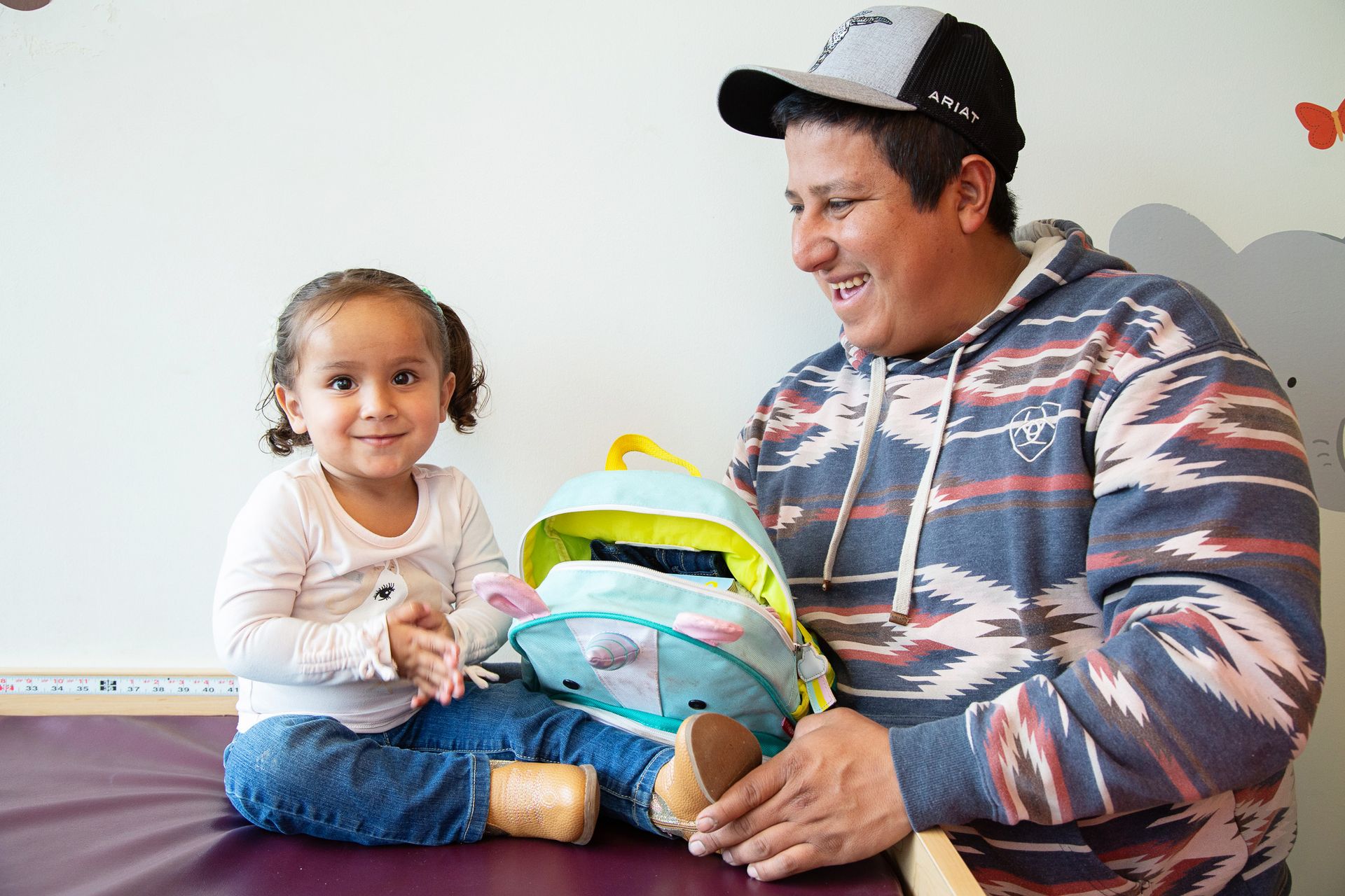 Smiling child with backpack next to smiling adult wearing a cap, in a light-colored room.